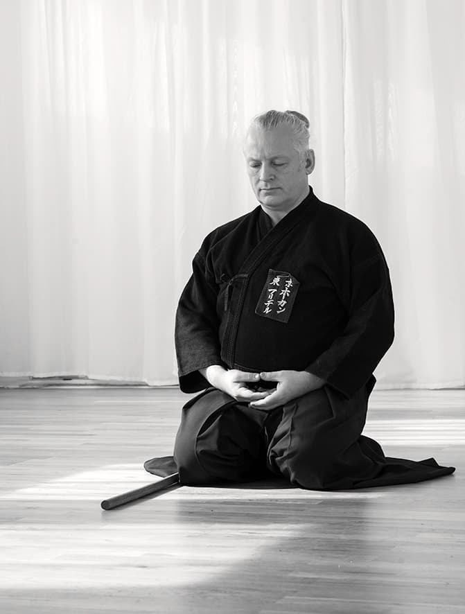 Håkan Friedel sitter i zazen i svart keikogi med ett träsvärd på golvet framför sig.